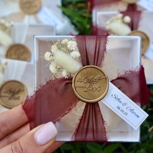 Op de afbeelding: Witte geschenkdozen met bordeauxrood lint, droogbloemen en een gouden zegel met de tekst "with love". Elke doos heeft een klein label met namen en een datum. De dozen staan op groen gras.