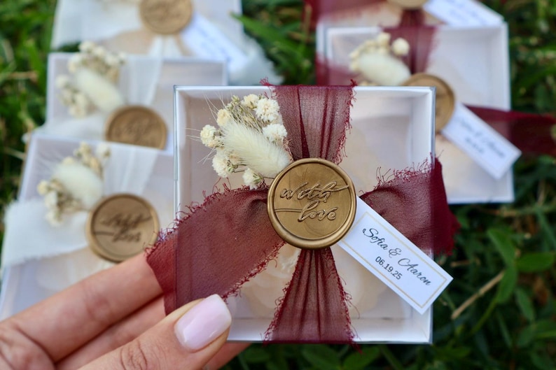 Op de afbeelding: Witte geschenkdozen met bordeauxrood lint, droogbloemen en een gouden zegel met de tekst "with love". Elke doos heeft een klein label met namen en een datum. De dozen staan op groen gras.