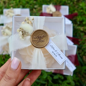 Op de afbeelding: Een transparante doos vastgebonden met een cr&egrave;mekleurig lint en een gouden zegel met de tekst "with love". De doos is versierd met gedroogde bloemen en een label met de tekst "Sofia & Aaron 06.19.25".