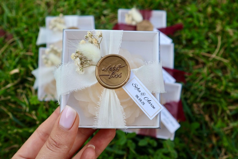 Op de afbeelding: Een transparante doos vastgebonden met een cr&egrave;mekleurig lint en een gouden zegel met de tekst "with love". De doos is versierd met gedroogde bloemen en een label met de tekst "Sofia & Aaron 06.19.25".