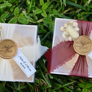 Op de afbeelding: Twee kleine, transparante dozen met deksel, elk versierd met een lint, gedroogde bloemen en een zegel met de tekst "sweet love". E&eacute;n doos heeft een cr&egrave;mekleurig lint, de andere een bordeauxrood lint. Elke doos heeft een label met "Sofia & Aaron 06.19.25."
