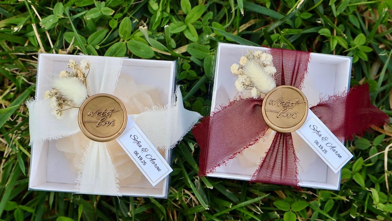 Op de afbeelding: Twee kleine, transparante dozen met deksel, elk versierd met een lint, gedroogde bloemen en een zegel met de tekst "sweet love". E&eacute;n doos heeft een cr&egrave;mekleurig lint, de andere een bordeauxrood lint. Elke doos heeft een label met "Sofia & Aaron 06.19.25."