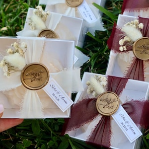 Op de afbeelding: Kleine, transparante doosjes, vastgebonden met ivoorkleurige of bordeauxrode linten, versierd met gedroogde bloemen en gouden waszegels. Elk doosje heeft een label met namen en een datum. De doosjes zijn gerangschikt op groen gras, wat duidt op een bruiloft of evenement.