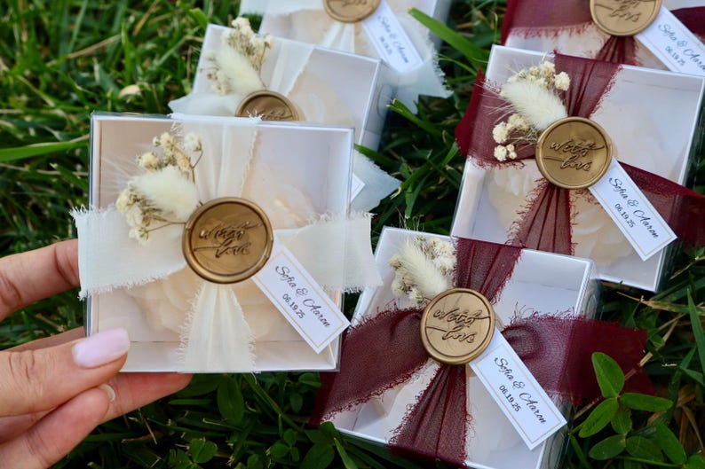Op de afbeelding: Kleine, transparante doosjes, vastgebonden met ivoorkleurige of bordeauxrode linten, versierd met gedroogde bloemen en gouden waszegels. Elk doosje heeft een label met namen en een datum. De doosjes zijn gerangschikt op groen gras, wat duidt op een bruiloft of evenement.