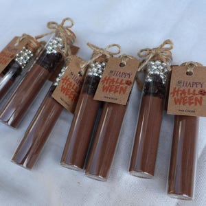 May include: Several glass tubes filled with brown powder, possibly hot cocoa mix, are arranged on a white surface. Each tube is sealed with a cork and tied with twine, and has a small tag that says "Happy Halloween" and "Hot Cocoa".