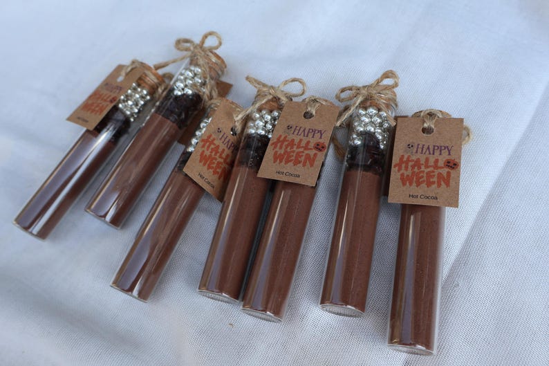 May include: Several glass tubes filled with brown powder, possibly hot cocoa mix, are arranged on a white surface. Each tube is sealed with a cork and tied with twine, and has a small tag that says "Happy Halloween" and "Hot Cocoa".