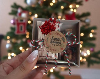 May include: A small gift box tied with red, green, and white twine, containing a small jar, a package, and dried red flowers. A round tag with "happy holidays" is attached. The background is a blurred Christmas tree with lights.