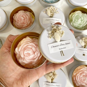 May include: A collection of floral-shaped candles in small, round, white tins with gold rims. Each candle is a different pastel color, including pink, blue, and white. The lids have a label that reads "Chama's Baby Shower 25.04.2023" and are decorated with dried flowers.