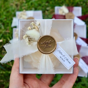 Op de afbeelding: Een witte vierkante geschenkdoos vastgebonden met een cr&egrave;mekleurig lint en een gouden waszegel met de tekst "with love". De doos is versierd met gedroogde bloemen en een label met de tekst "Sofia & Aaron 06.19.25."