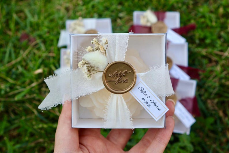 Op de afbeelding: Een witte vierkante geschenkdoos vastgebonden met een cr&egrave;mekleurig lint en een gouden waszegel met de tekst "with love". De doos is versierd met gedroogde bloemen en een label met de tekst "Sofia & Aaron 06.19.25."
