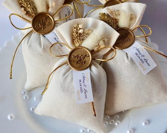 May include: White fabric favor bags with gold ribbon and a gold wax seal. Each bag has a small tag with the text "Ashley & Lucas 06.14.2025".
