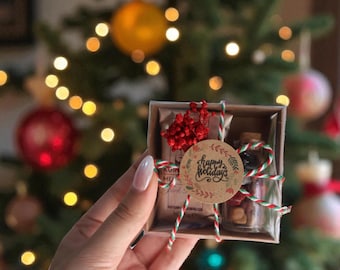 May include: A small gift box with a festive theme, featuring a round label that reads "happy holidays." The box contains small glass bottles and decorative elements, tied with red and green twine. The background includes a Christmas tree with ornaments and lights.