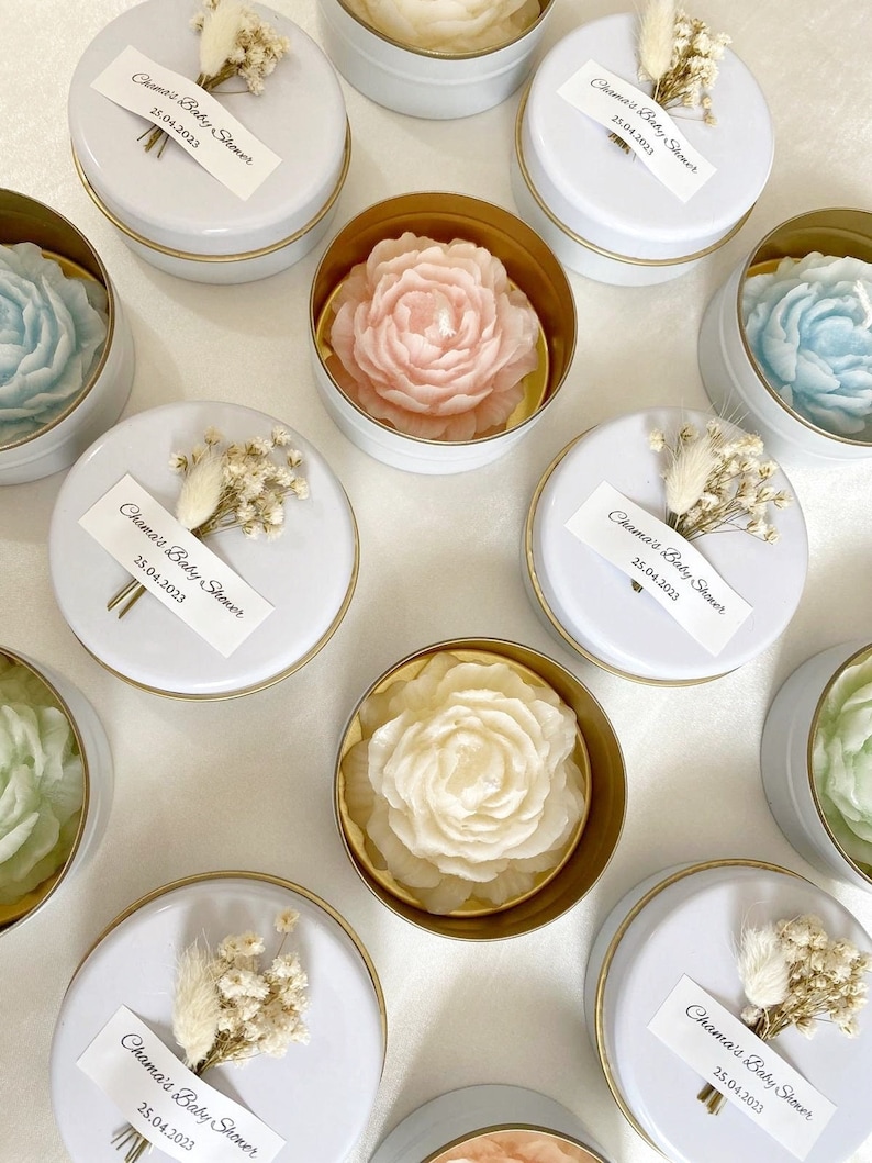 May include: A collection of floral-shaped candles in small, round, white tins with gold rims. The candles are various pastel colors, including pink, blue, and green. Each tin is topped with a white label that reads "Chama's Baby Shower 25.04.2023" and a small floral arrangement.