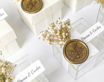 May include: White favor boxes with a gold wax seal that says "with love" and a white label with the text "Denise & Carlos". The boxes are decorated with dried white flowers.