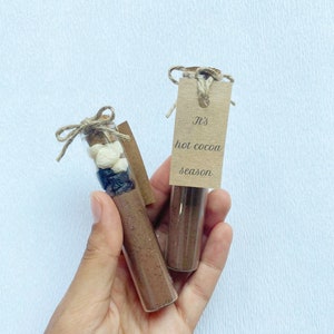 May include: Two glass test tubes filled with hot cocoa mix, marshmallows, and chocolate chips. Each tube has a brown paper tag tied around it with twine. The tag on the right tube reads "It's hot cocoa season."