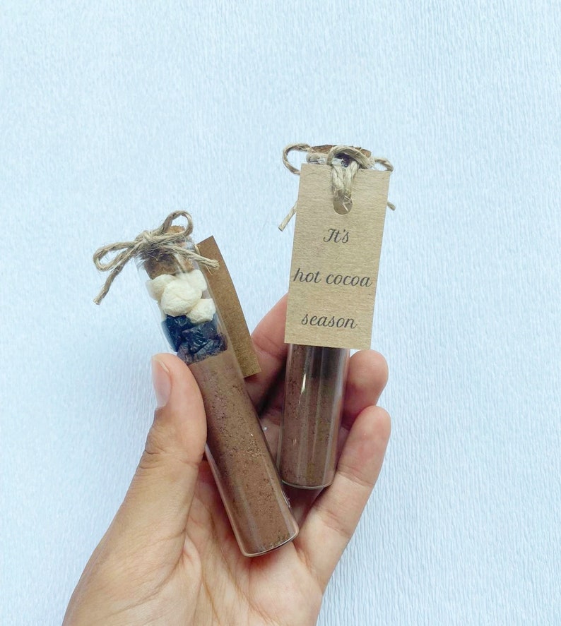 May include: Two glass test tubes filled with hot cocoa mix, marshmallows, and chocolate chips. Each tube has a brown paper tag tied around it with twine. The tag on the right tube reads "It's hot cocoa season."