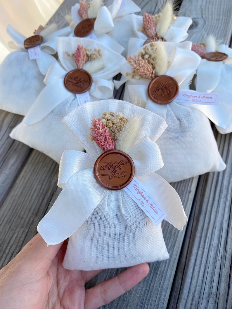 Puede incluir: Bolsas de tela blanca con un sello de cera marr&oacute;n y flores secas. Las bolsas tienen una cinta atada alrededor y una etiqueta que dice "Meghan & Adam 5.25.21".