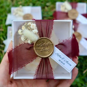 Op de afbeelding: Een transparante vierkante doos vastgebonden met een bordeauxrood lint en een gouden zegel met de tekst "with love". De doos bevat een klein wit object en is versierd met gedroogde bloemen. Een klein label leest "Sofia & Aaron 06.19.25".