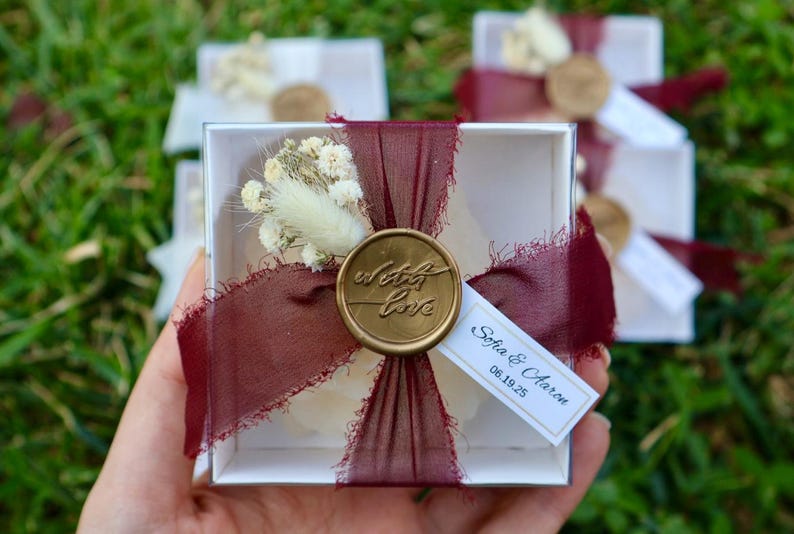 Op de afbeelding: Een transparante vierkante doos vastgebonden met een bordeauxrood lint en een gouden zegel met de tekst "with love". De doos bevat een klein wit object en is versierd met gedroogde bloemen. Een klein label leest "Sofia & Aaron 06.19.25".