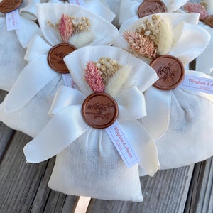 Puede incluir: Bolsas de tela blanca con un sello de cera marr&oacute;n y flores secas. Las bolsas tienen una cinta blanca y una etiqueta que dice "Meghan & Adam 5/25/24".