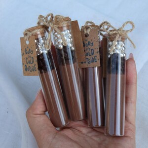 May include: Several glass tubes filled with a brown liquid and topped with corks and decorative silver beads. Each tube has a small tag with the text "Baby it's Cold Outside" tied with twine. The tubes are held in a hand.