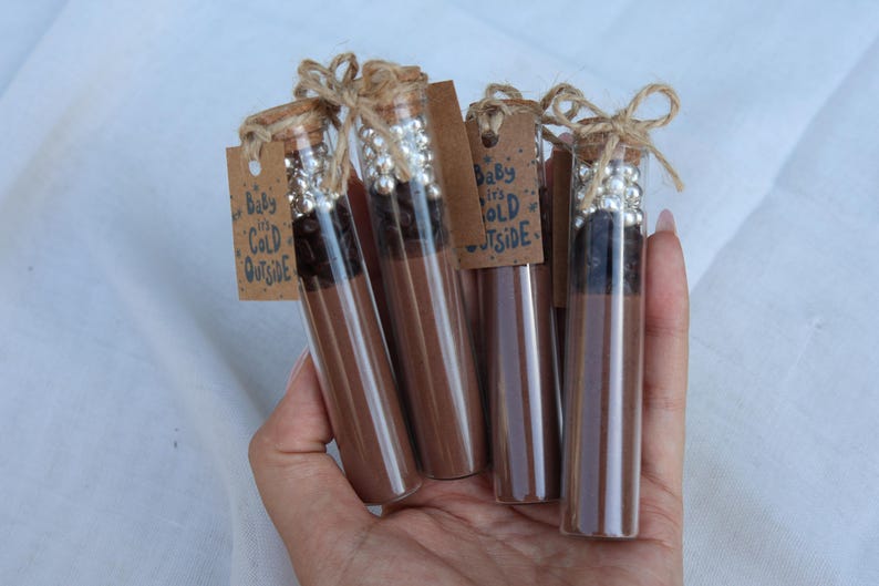 May include: Several glass tubes filled with a brown liquid and topped with corks and decorative silver beads. Each tube has a small tag with the text "Baby it's Cold Outside" tied with twine. The tubes are held in a hand.