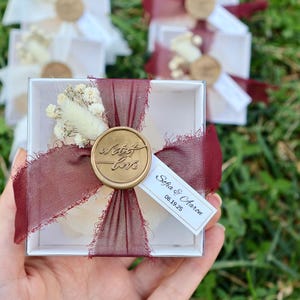 Op de afbeelding: Een kleine witte geschenkdoos, vastgebonden met een bordeauxrood lint en verzegeld met een gouden wasstempel met de tekst "With Love". De doos is versierd met gedroogde bloemen en een klein label met namen en een datum.