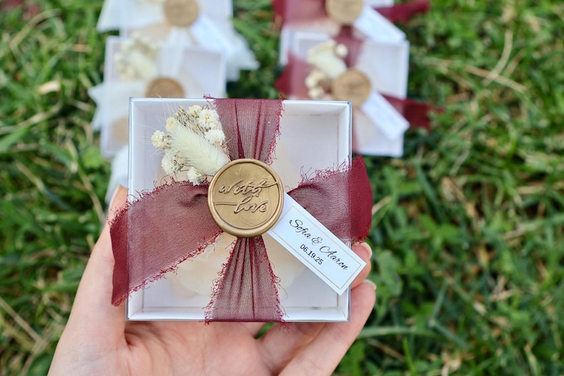 Op de afbeelding: Een kleine witte geschenkdoos, vastgebonden met een bordeauxrood lint en verzegeld met een gouden wasstempel met de tekst "With Love". De doos is versierd met gedroogde bloemen en een klein label met namen en een datum.