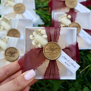 Op de afbeelding: Kleine witte geschenkdozen, vastgebonden met bordeauxrood lint en verzegeld met een gouden wasstempel met de tekst "with love". Elke doos is versierd met gedroogde bloemen en een klein label met namen en een datum. De dozen zijn gerangschikt op groen gras.