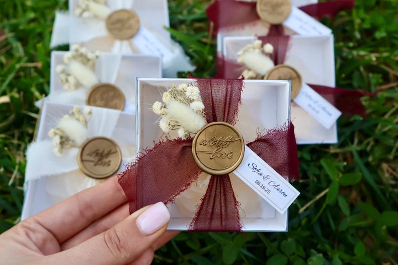 Op de afbeelding: Kleine witte geschenkdozen, vastgebonden met bordeauxrood lint en verzegeld met een gouden wasstempel met de tekst "with love". Elke doos is versierd met gedroogde bloemen en een klein label met namen en een datum. De dozen zijn gerangschikt op groen gras.
