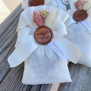 Puede incluir: Bolsas de tela blanca con un sello de cera marr&oacute;n que dice "with love" y flores secas. Las bolsas tienen una cinta blanca atada alrededor y una etiqueta blanca que dice "Meghan & Adam" con la fecha "5/25/24".