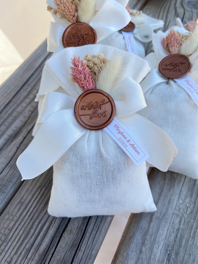 Puede incluir: Bolsas de tela blanca con un sello de cera marr&oacute;n que dice "with love" y flores secas. Las bolsas tienen una cinta blanca atada alrededor y una etiqueta blanca que dice "Meghan & Adam" con la fecha "5/25/24".