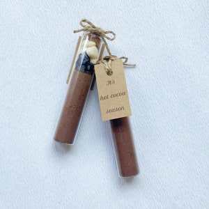 May include: Two glass test tubes filled with brown powder and topped with a mixture of dried fruit and nuts. A brown paper tag is tied to one of the tubes with the text "It's hot cocoa season".