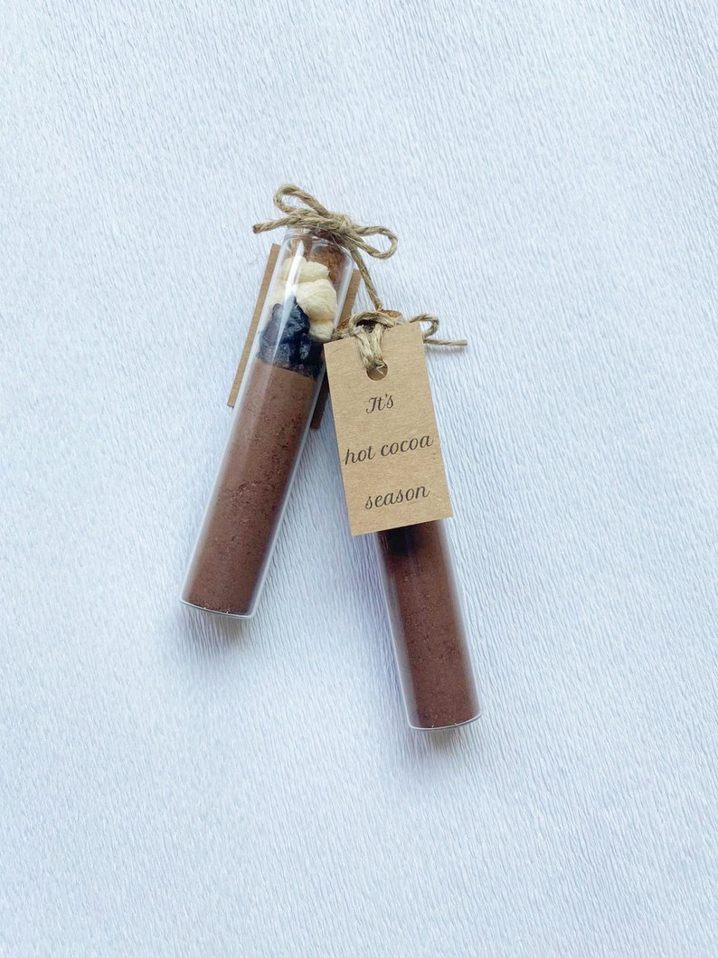 May include: Two glass test tubes filled with brown powder and topped with a mixture of dried fruit and nuts. A brown paper tag is tied to one of the tubes with the text "It's hot cocoa season".