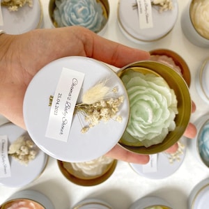 May include: A collection of small, round tins with gold rims and white lids, each containing a flower-shaped candle. The candles are in various pastel colors, including blue, green, and cream. Each tin is decorated with a small sprig of dried flowers and a label that reads "Chama's Baby Shower 25.04.2023".