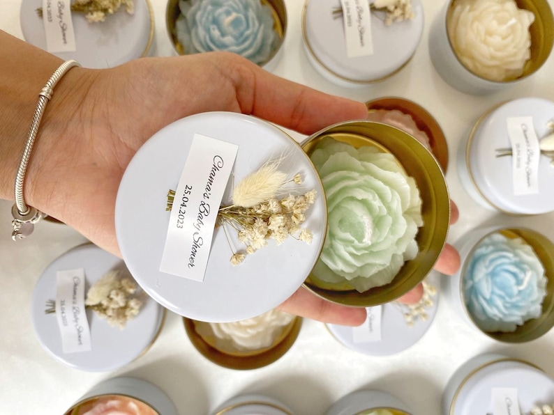 May include: A collection of small, round tins with gold rims and white lids, each containing a flower-shaped candle. The candles are in various pastel colors, including blue, green, and cream. Each tin is decorated with a small sprig of dried flowers and a label that reads "Chama's Baby Shower 25.04.2023".
