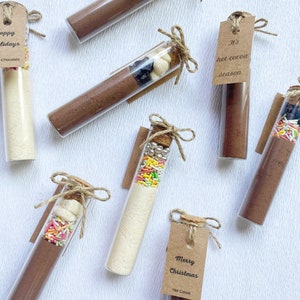 May include: Assortment of glass test tubes filled with hot cocoa mix, marshmallows, sprinkles, and other toppings. Each tube is tied with twine and a brown paper tag with a festive message. The tags read "It's hot cocoa season", "Happy Holidays", "Merry Christmas", and "Chris & Emily".