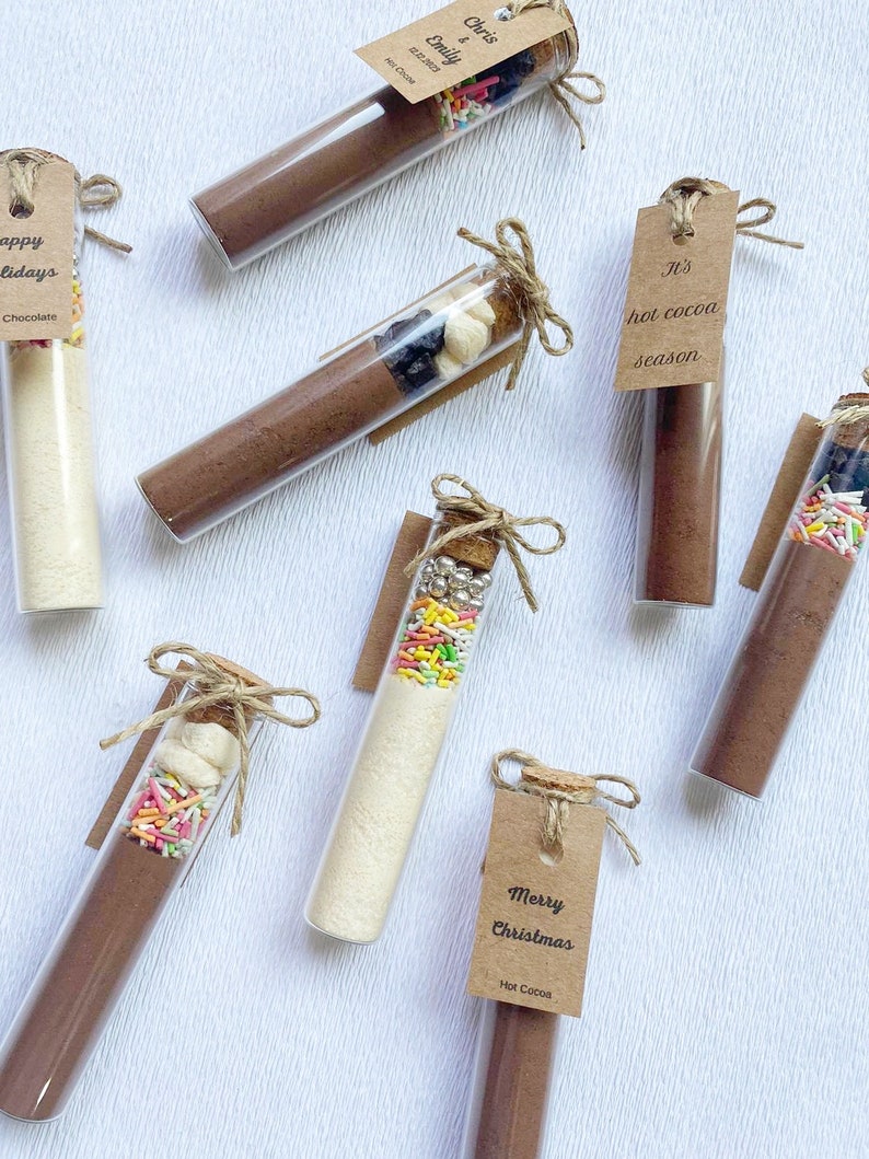 May include: Assortment of glass test tubes filled with hot cocoa mix, marshmallows, sprinkles, and other toppings. Each tube is tied with twine and a brown paper tag with a festive message. The tags read "It's hot cocoa season", "Happy Holidays", "Merry Christmas", and "Chris & Emily".