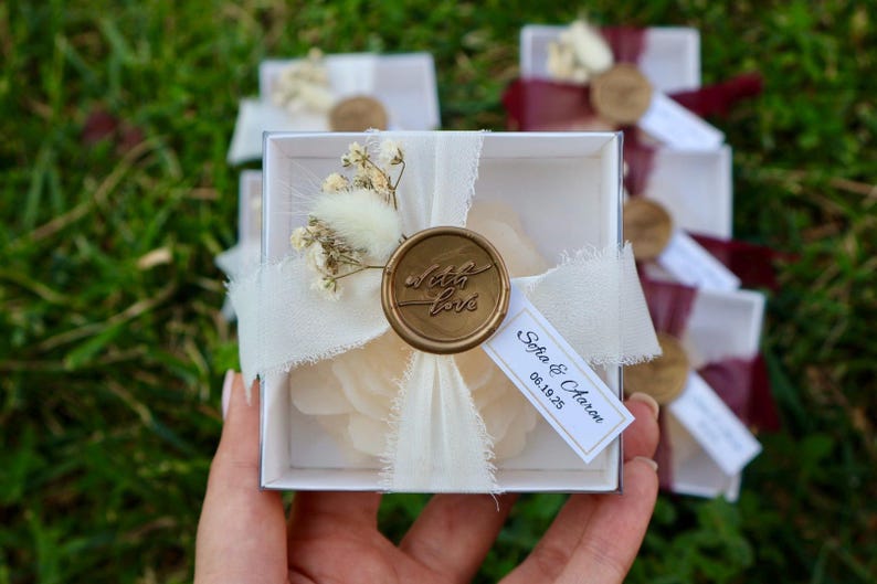 Op de afbeelding: Een witte geschenkdoos met een cr&egrave;mekleurige bloem, verzegeld met een gouden wasstempel met de tekst "with love". De doos is vastgebonden met een wit lint en een klein label met namen en een datum. Andere dozen zijn zichtbaar op de achtergrond.