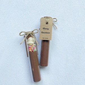 May include: Two glass test tubes filled with hot cocoa mix and toppings. One tube has marshmallows, sprinkles, and cocoa powder. The other tube has only cocoa powder. Both tubes have a brown paper tag that says "Merry Christmas" and "Hot Cocoa".