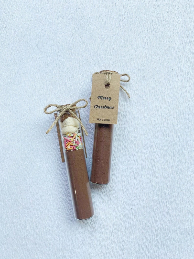 May include: Two glass test tubes filled with hot cocoa mix and toppings. One tube has marshmallows, sprinkles, and cocoa powder. The other tube has only cocoa powder. Both tubes have a brown paper tag that says "Merry Christmas" and "Hot Cocoa".