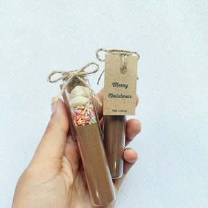 May include: Two small glass jars with brown lids and twine bows. The jars are filled with brown powder, white marshmallows, and colorful sprinkles. A brown paper tag is attached to one jar with the text "Merry Christmas Hot Cocoa".