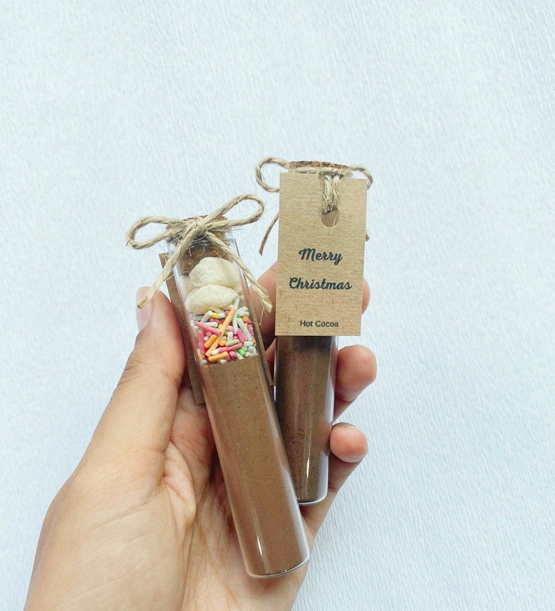 May include: Two small glass jars with brown lids and twine bows. The jars are filled with brown powder, white marshmallows, and colorful sprinkles. A brown paper tag is attached to one jar with the text "Merry Christmas Hot Cocoa".