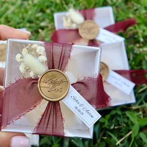 Op de afbeelding: Kleine, transparante doosjes vastgebonden met bordeauxrood lint en versierd met een gouden waszegel met de tekst "with love". Elk doosje bevat gedroogde bloemen en een klein label met namen en een datum.