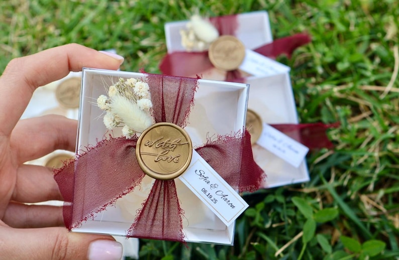 Op de afbeelding: Kleine, transparante doosjes vastgebonden met bordeauxrood lint en versierd met een gouden waszegel met de tekst "with love". Elk doosje bevat gedroogde bloemen en een klein label met namen en een datum.