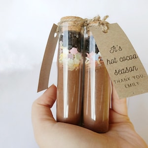 May include: Two glass test tubes filled with hot cocoa mix and topped with sprinkles. A brown paper tag is tied around the tubes with the text "It's hot cocoa season. Thank you. Emily."