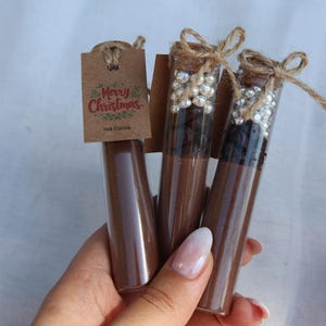 May include: Three glass tubes filled with brown powder, possibly hot cocoa mix, are displayed. Two tubes have decorative pearl-like beads and are sealed with corks and twine. A small tag reads "Merry Christmas Hot Cocoa."