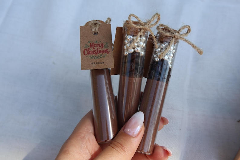 May include: Three glass tubes filled with brown powder, possibly hot cocoa mix, are displayed. Two tubes have decorative pearl-like beads and are sealed with corks and twine. A small tag reads "Merry Christmas Hot Cocoa."