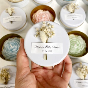 May include: Baby shower favors featuring rose-shaped soaps in various pastel colors, including white, pink, blue, and green. Each soap is nestled in a small, round, white tin with a gold rim, adorned with dried flowers and a label that reads "Chama's Baby Shower 25.04.2023."