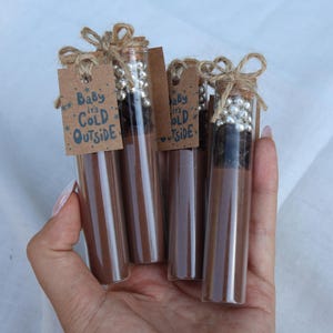 May include: A hand holds four glass tubes filled with a brown substance, each capped with silver beads and tied with twine. Small brown tags read "Baby it's Cold Outside". The tubes are a winter-themed gift.